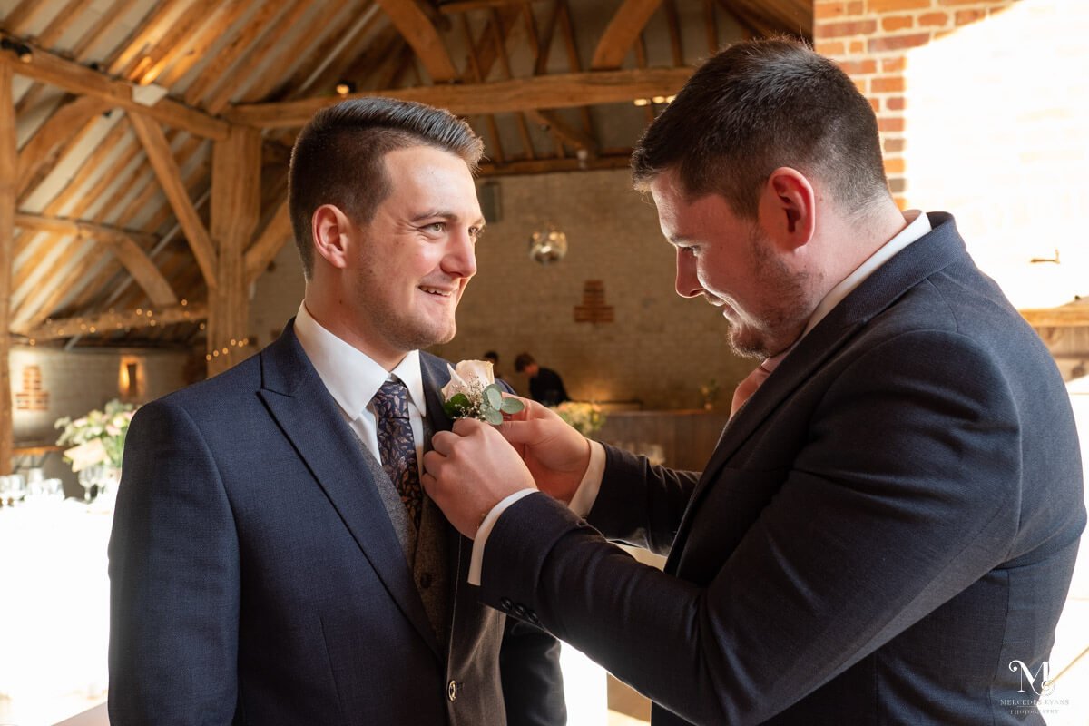 the groomsnman pins on the grooms buttonhole in the barn at Bury Court