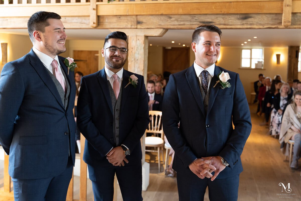 the groom smiles across the room as he waits with his two best men for the bridal party to arrive