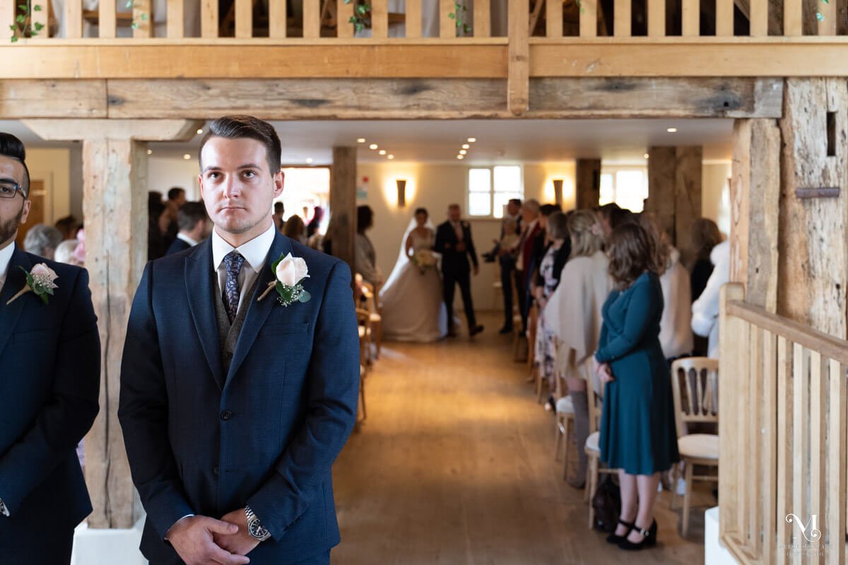 the anxious groom looks up at the barn roof and behind him the bride is walking down the aisle with her father