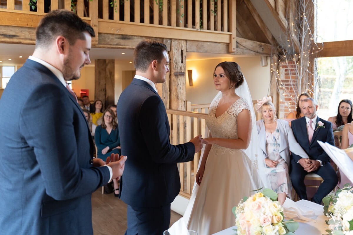 The groom puts the wedding ring on to his bride's finger, the best man stands behind holding the other ring and her parents watch from behind her