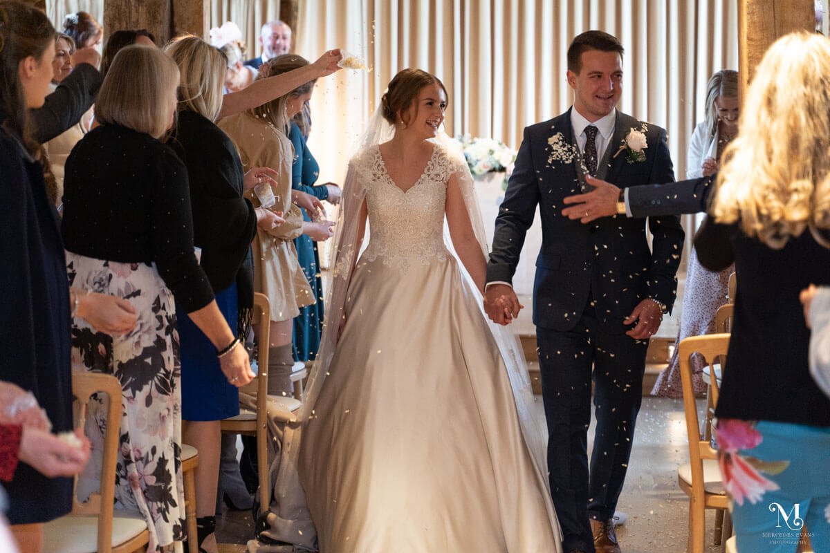 the bride and groom walk down the aisle hand in hand and the guests throw confetti at the barn at Bury Court