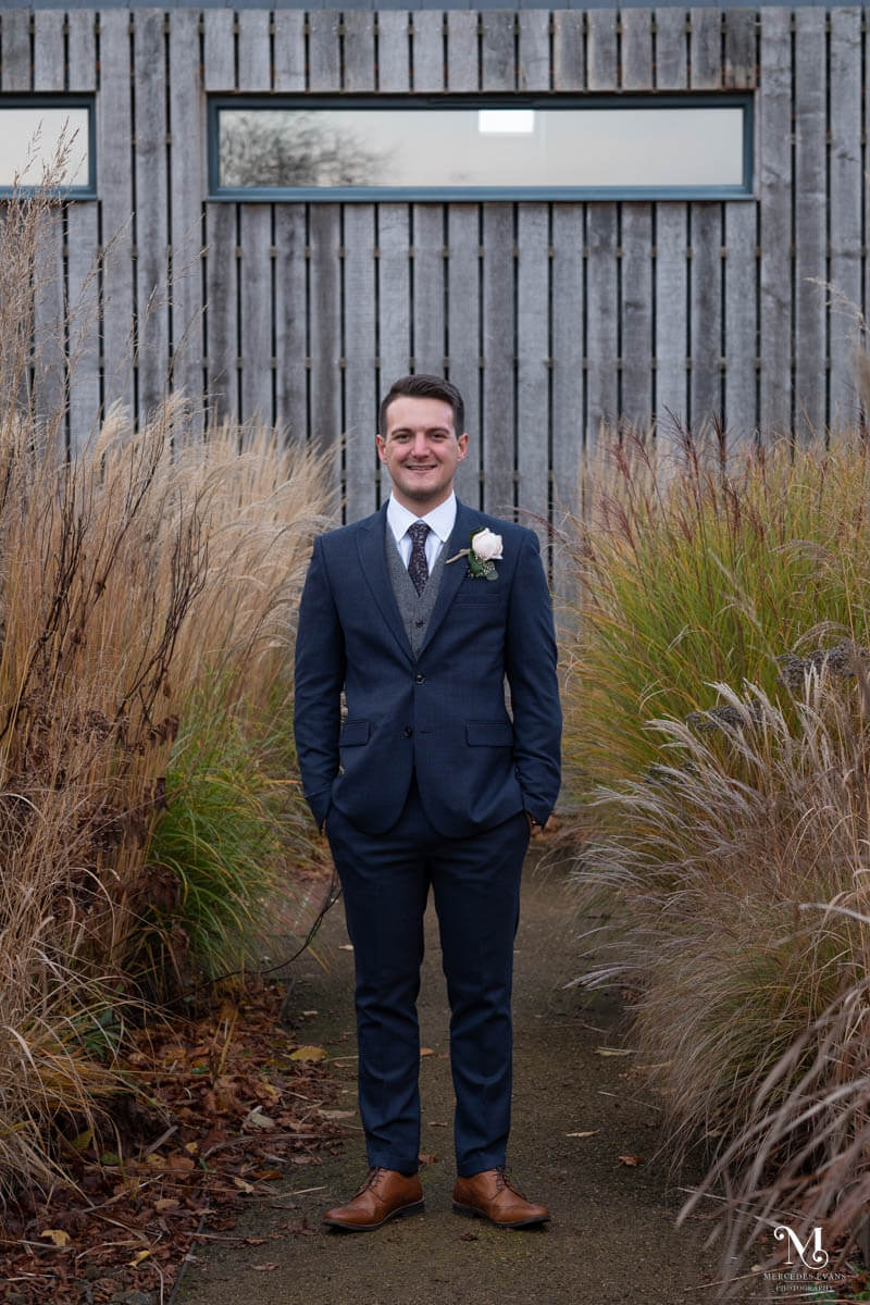 the smiling groom stands square to the camera with both hands in his trouser pockets. He is in front of a wood panelled building with long grasses on either side of him