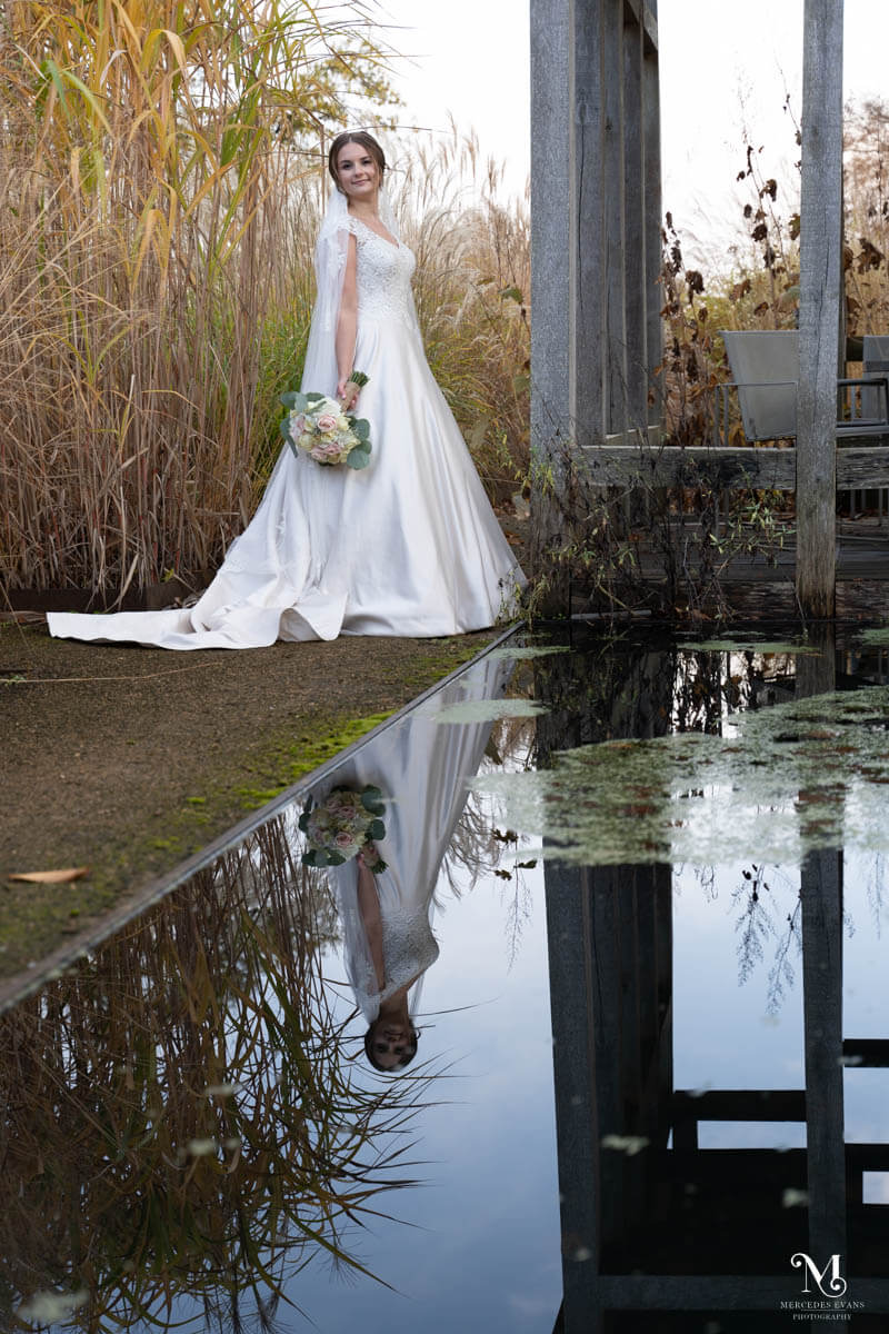 the bride looks back over her shoulder, she is between a metal structure and long grasses and is reflected in the pool in front of her