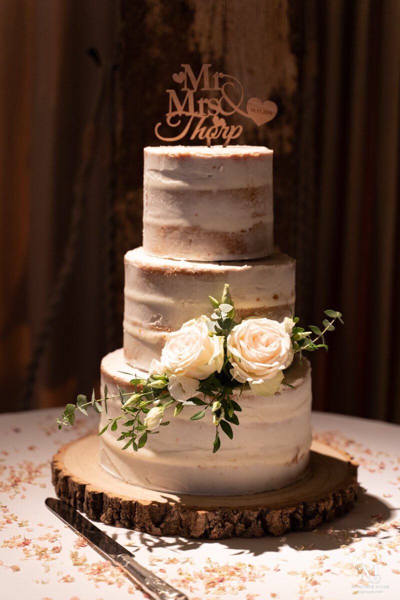 a three tiered semi-iced wedding cake with a wooden topper, white roses and eucalyptus