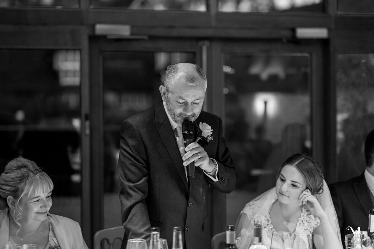 black and white of the father of the bride during his speech, his wife and the bride sit on either side of him