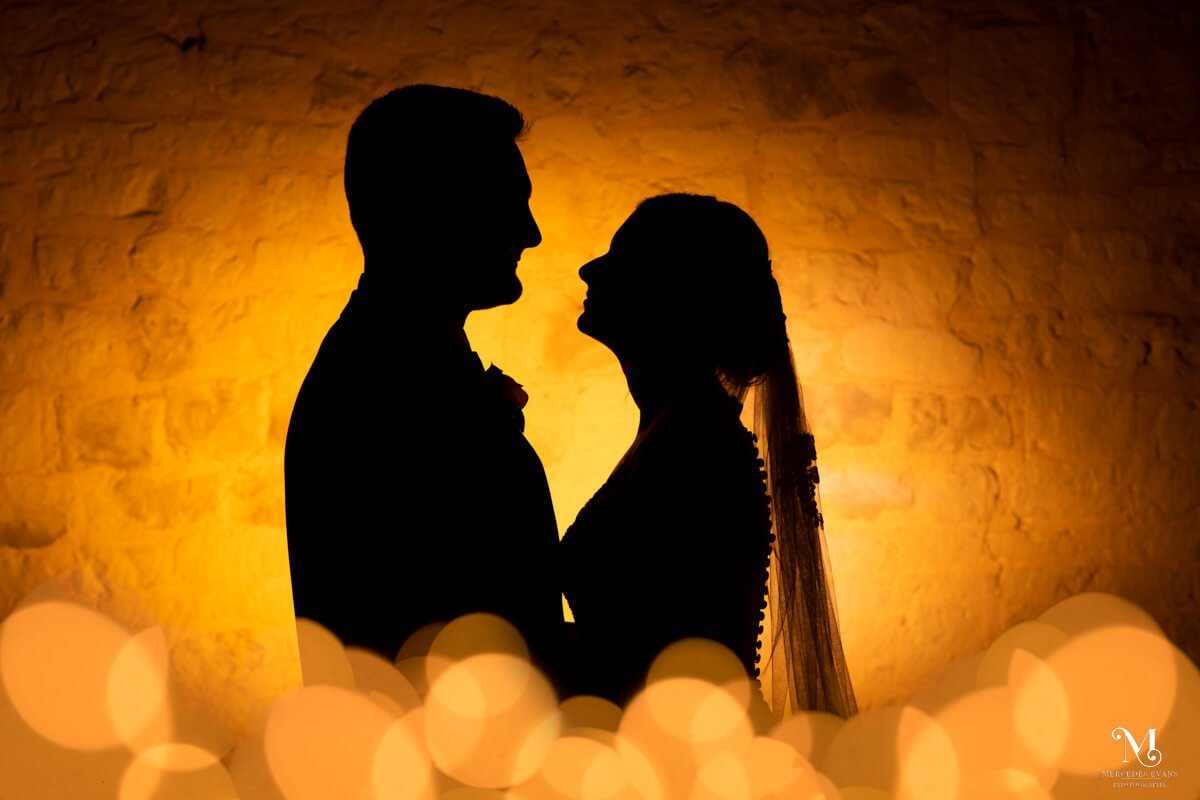 a silhouette of the bride and groom against a yellow wall with fairy lights at the bottom of the image
