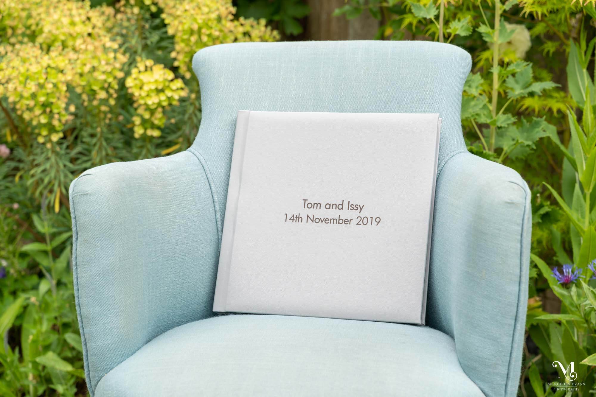 a pale grey wedding album with the words tom and issy 14th november 2019, sits on a pale blue arm chair in a garden with flowers