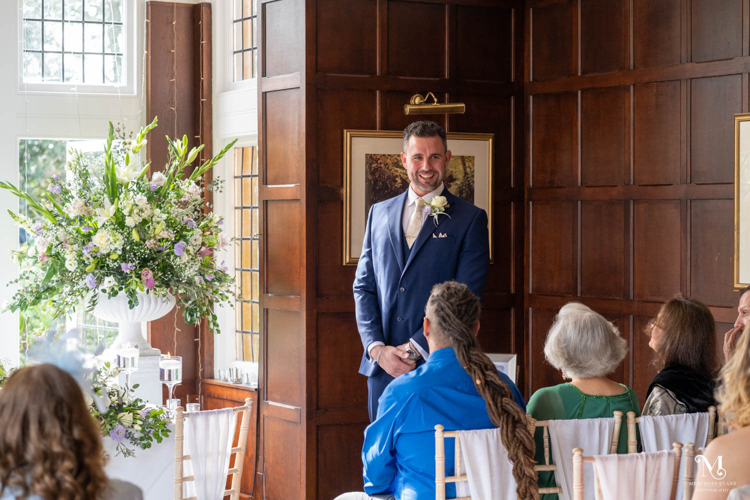 the groom stands in front of guests as they wait for the bridal party in the Oak room at Cantley House