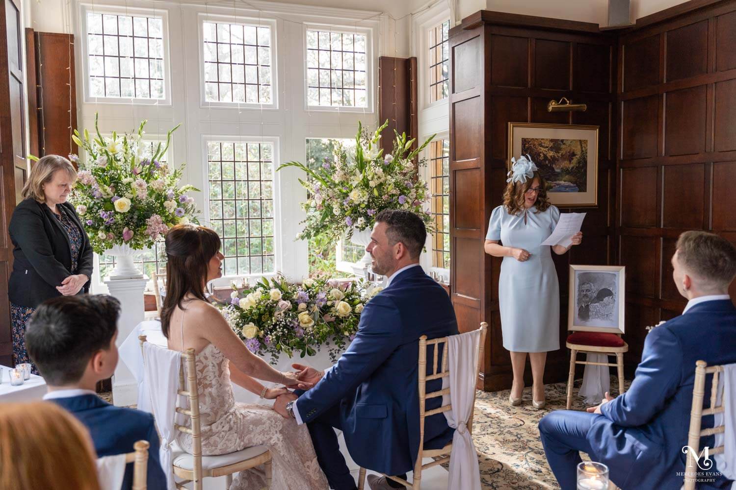 The bride and groom look at each other and hold hands as a female guest does a reading in the Oak room at Cantley House