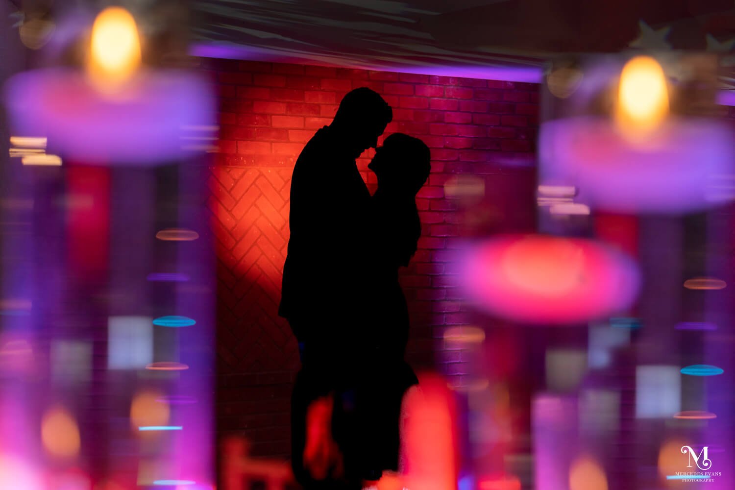 the bride and groom are silhouetted against a red lit wall and in the foreground are red, blue, purple and gold shapes