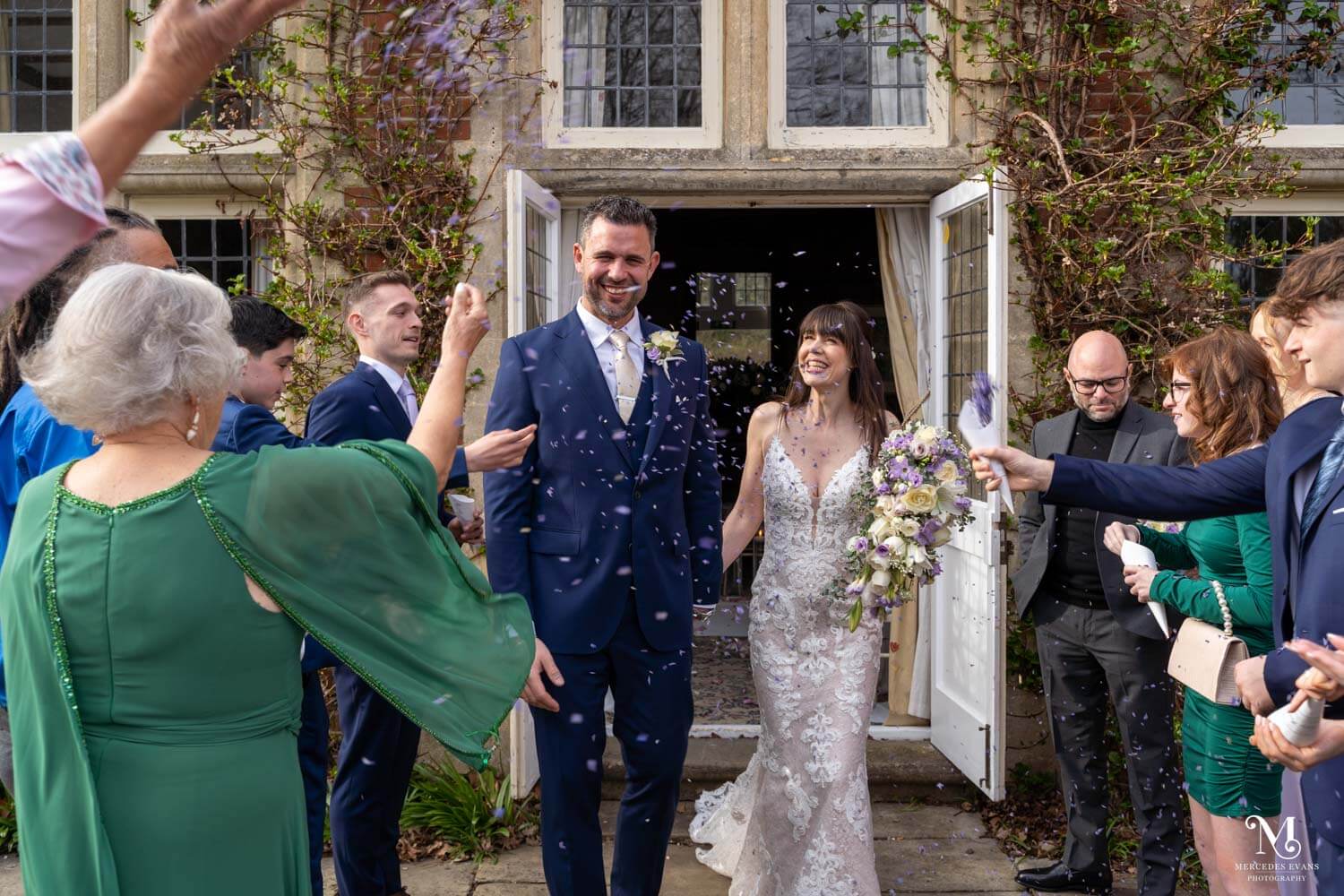 the bride and groom walk hand in hand as guests throw lilac confetti outside the Oak room at Cantley House Hotel
