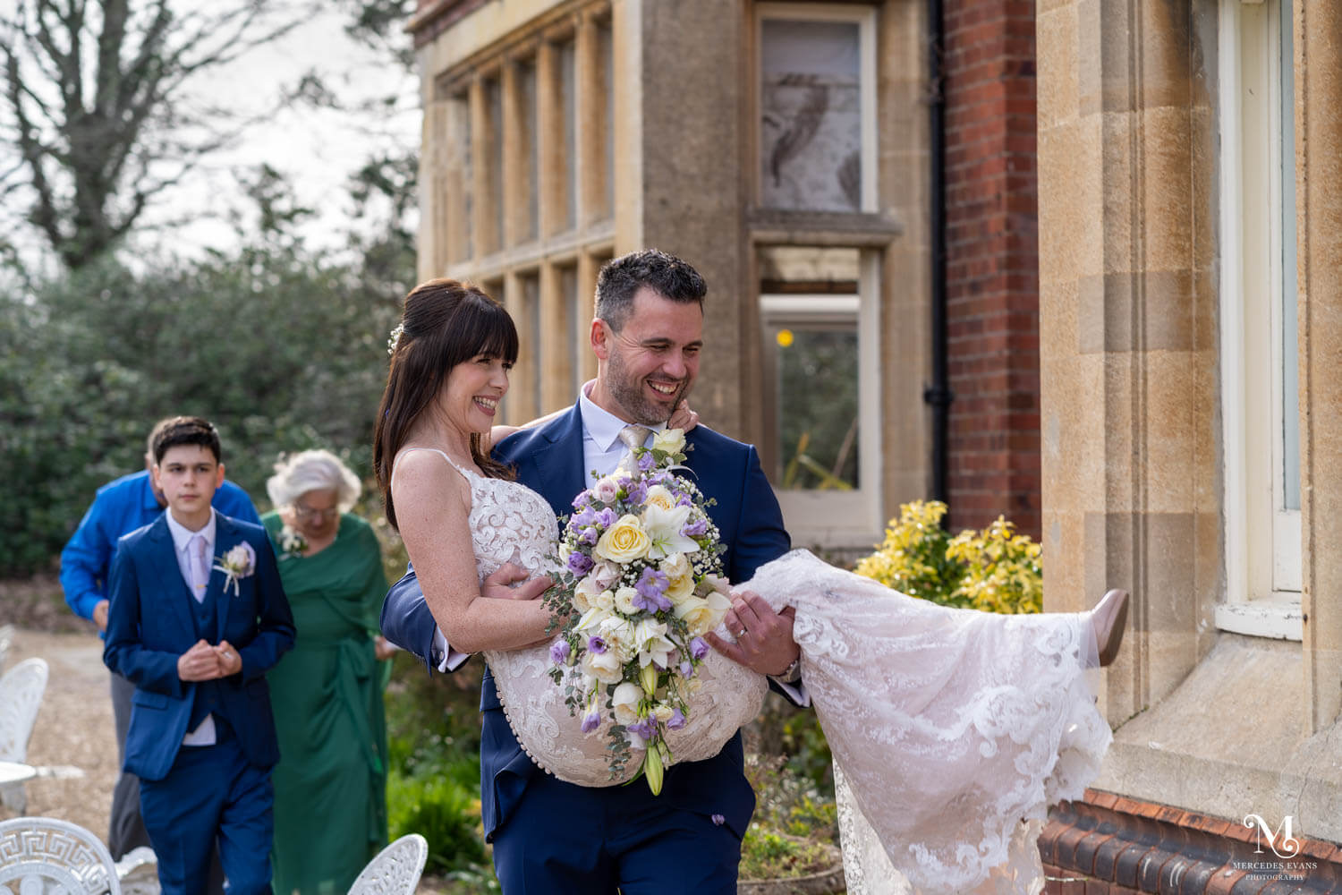 the groom carries his bride, surrounded by wedding guests as they walk in the garden at Cantley house