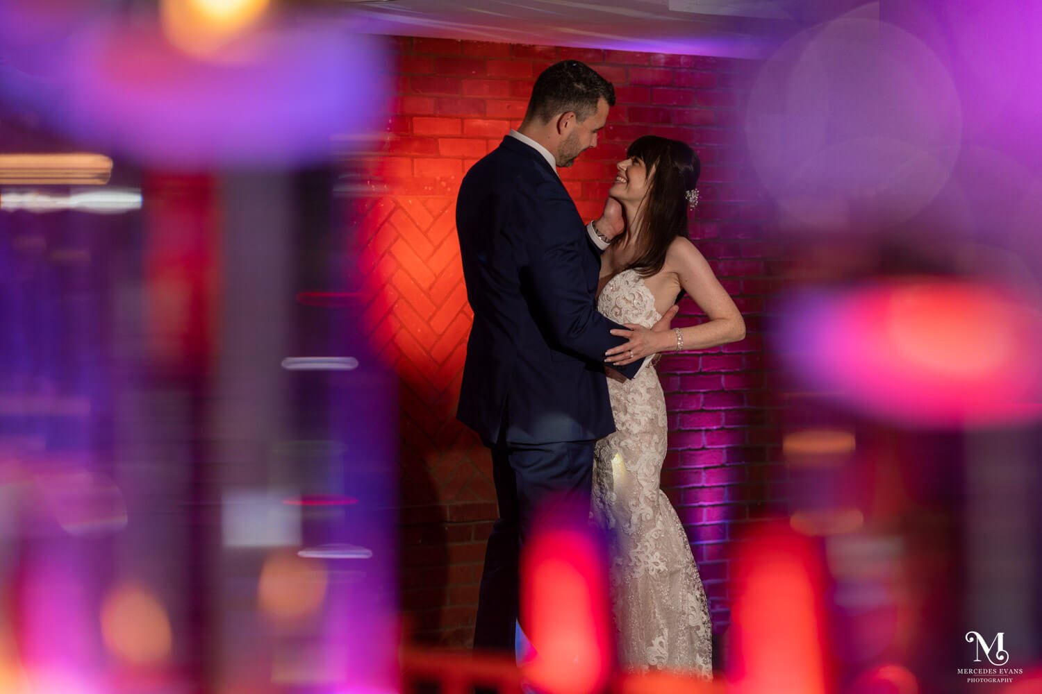an evening photo of the bride and groom laughing as they embrace, with purple and red lights in the foreground