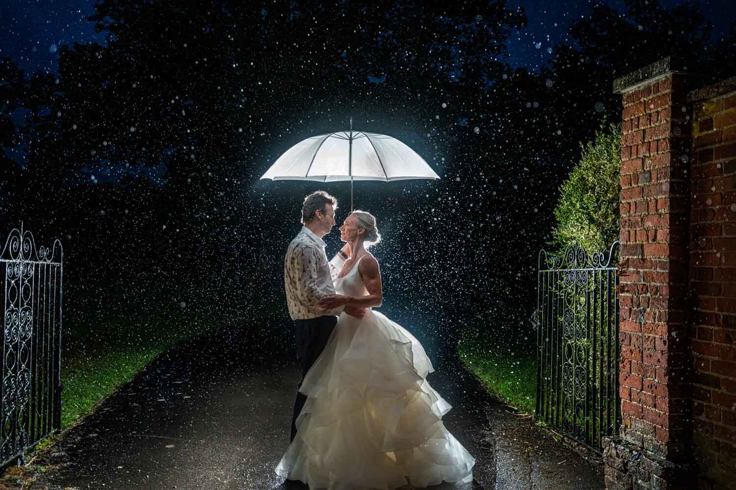 bride and groom embrace under an umbrella in the rain at night time and they are standing in the gateway at Warbrook house