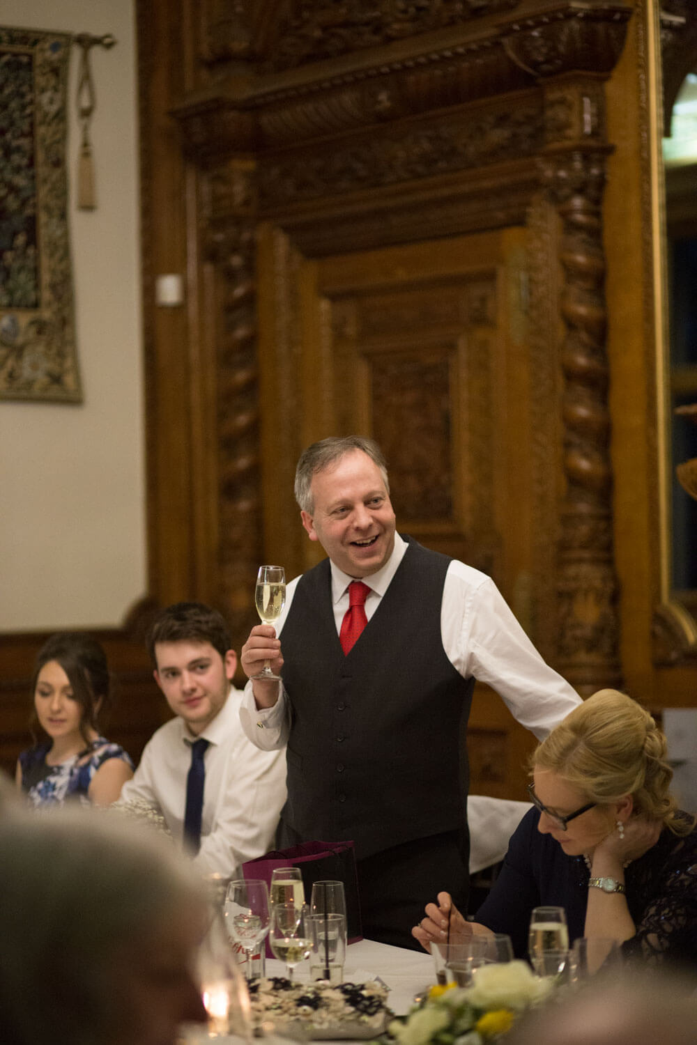 smiling man stands holding a glass of champagne as he makes a speech at the wedding breakfast table