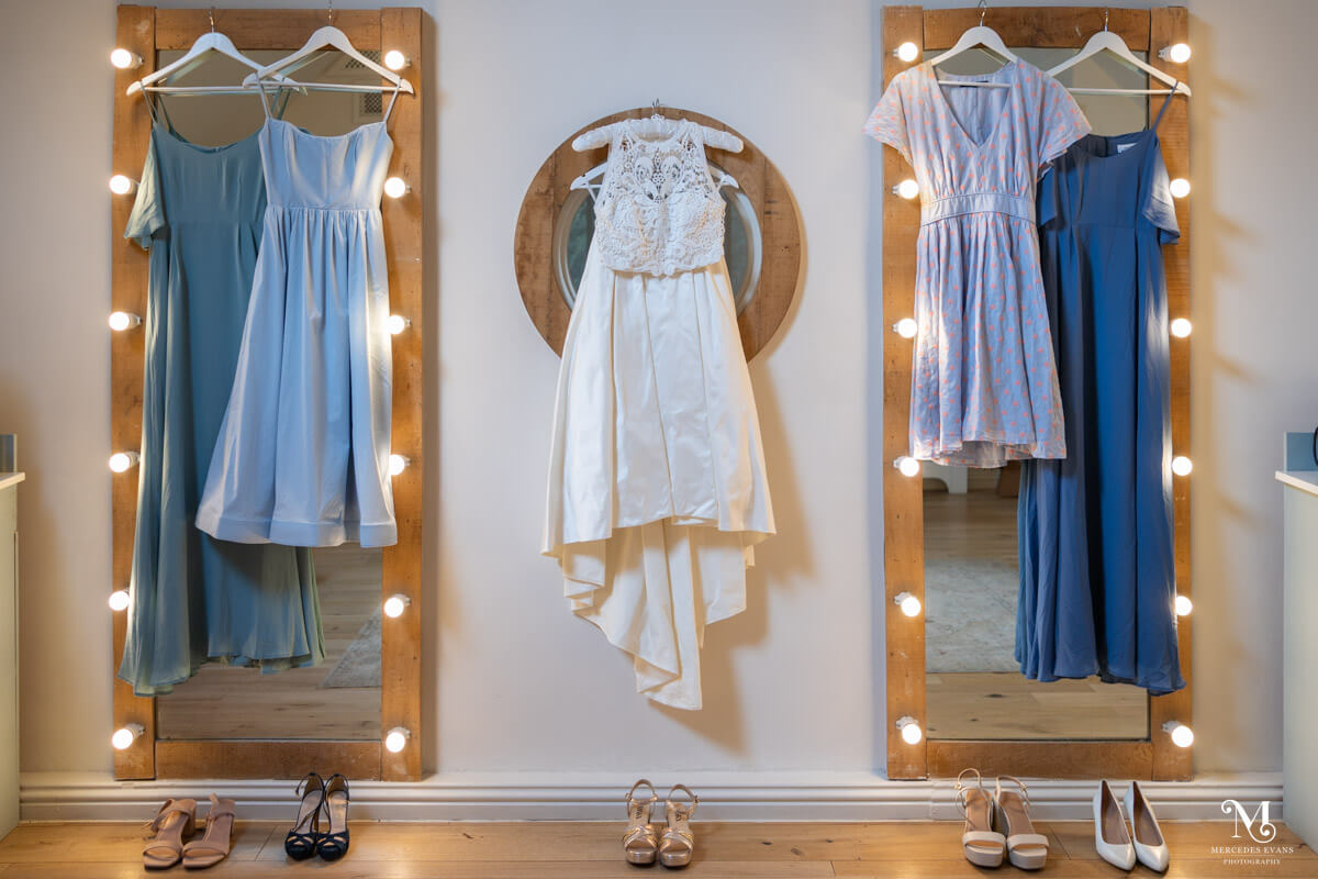 two different blue bridesmaids dresses hang on hollywood mirrors either side of the bride's dress, the wedding shoes are underneath each of the five dresses