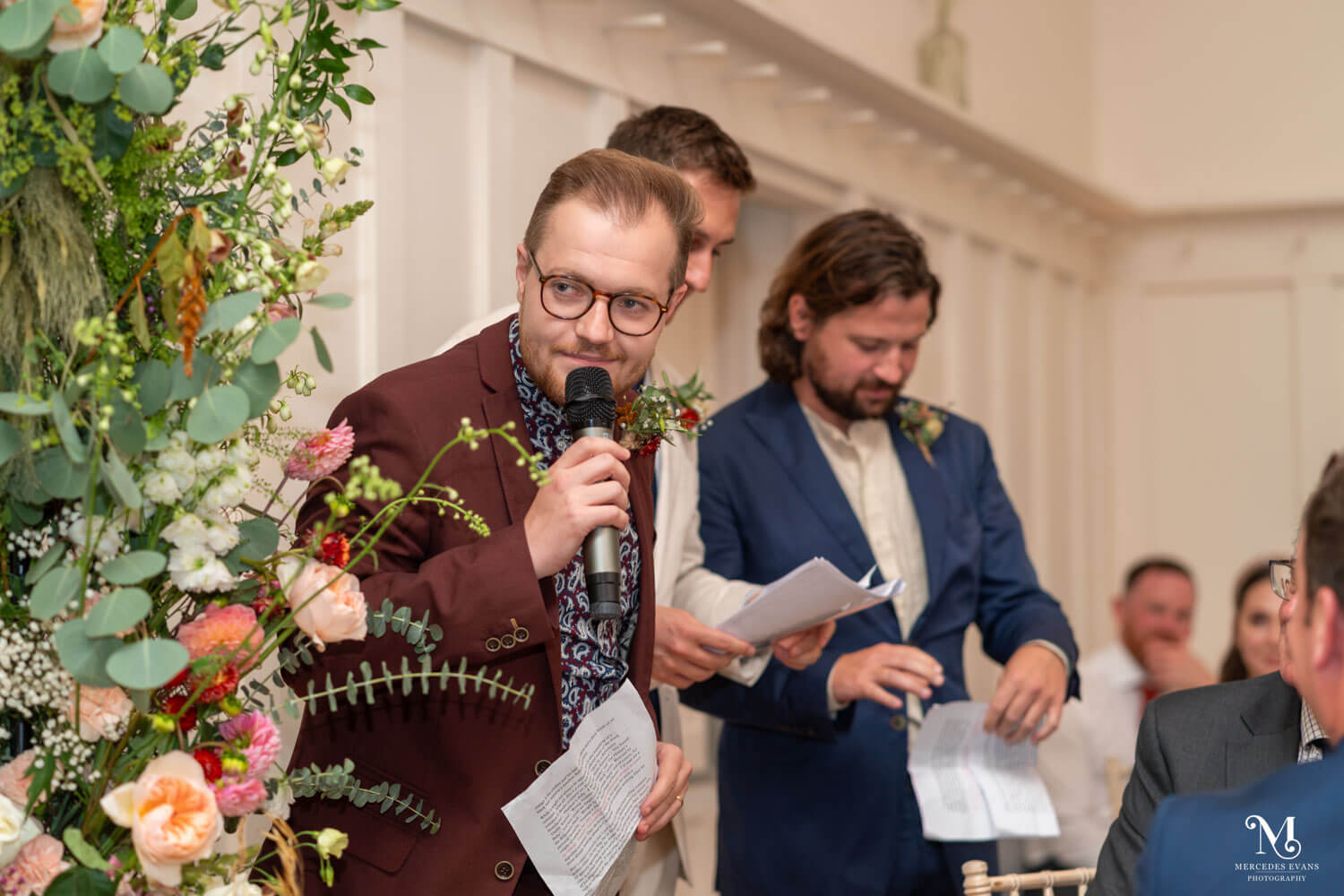 three bestmen are making their speeches, the closest holds the microphone and peers round the foliage, the other two and looking at their papers