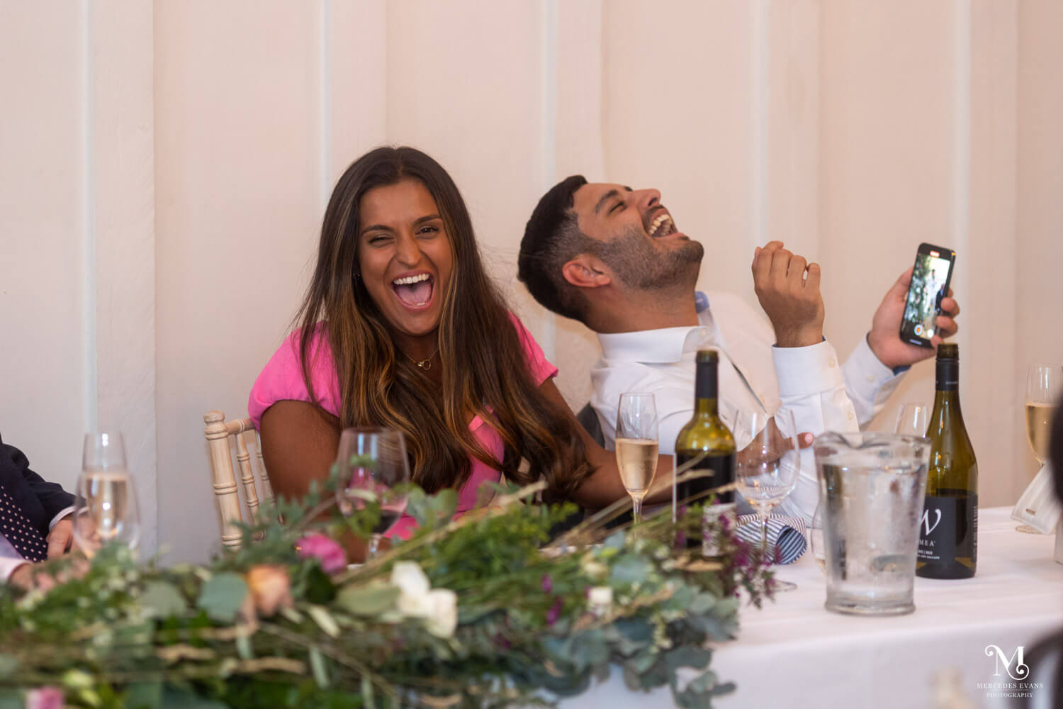 a man and woman laugh hysterically, the man holds his phone which he is using to film the bestman's speech
