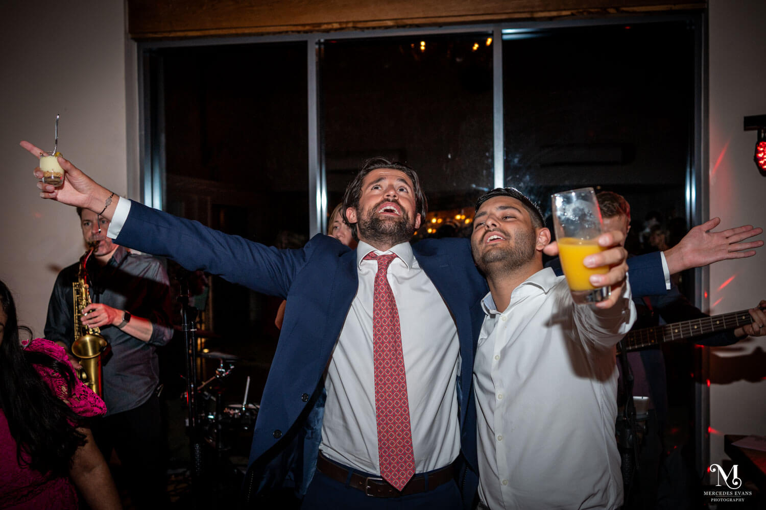 two male guests sing, with drinks and pudding in hand and arms outstretched on the dance floor