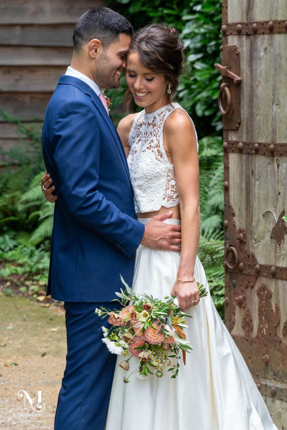 the smiling groom holds his bride by the waist as she looks down at her wedding bouquet, they stand at the gateway to the Hideaway garden at millbridge court