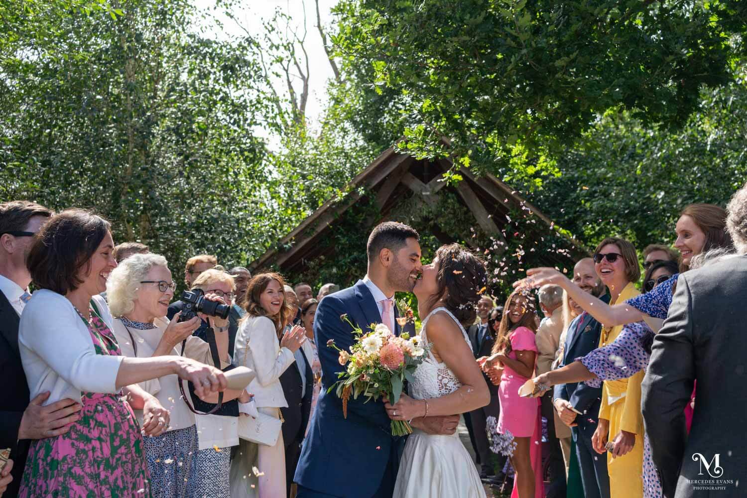 bride and groom kiss and laugh as guests throw confetti by the oak arbour at millbridge court
