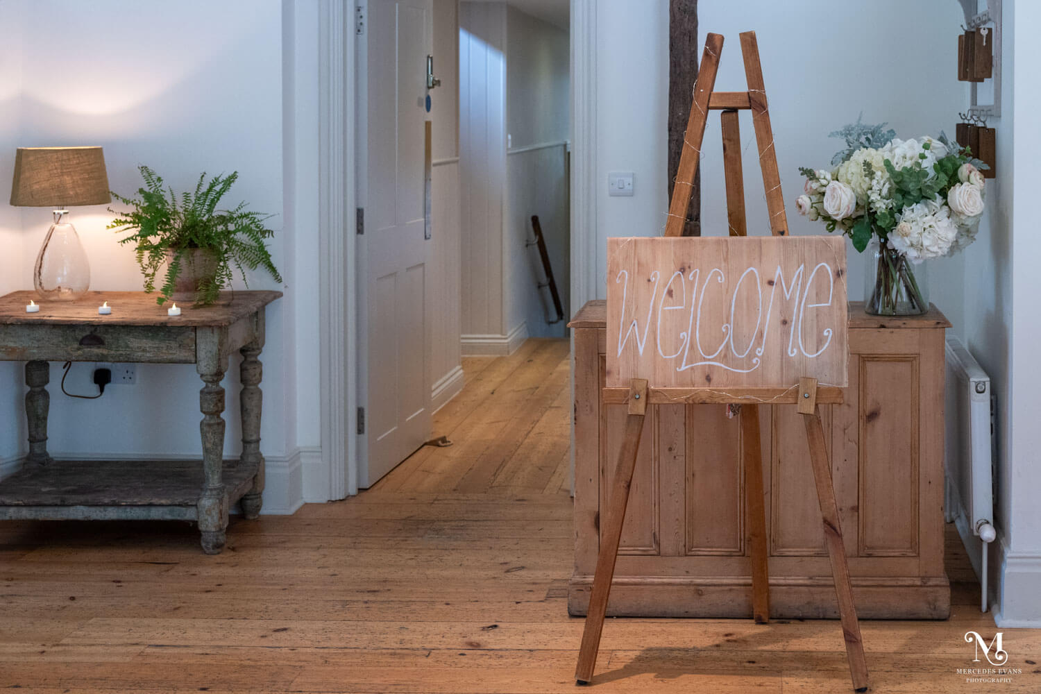 a wooden welcome sign sits on an easel, behind this is a large vase of white flowers.