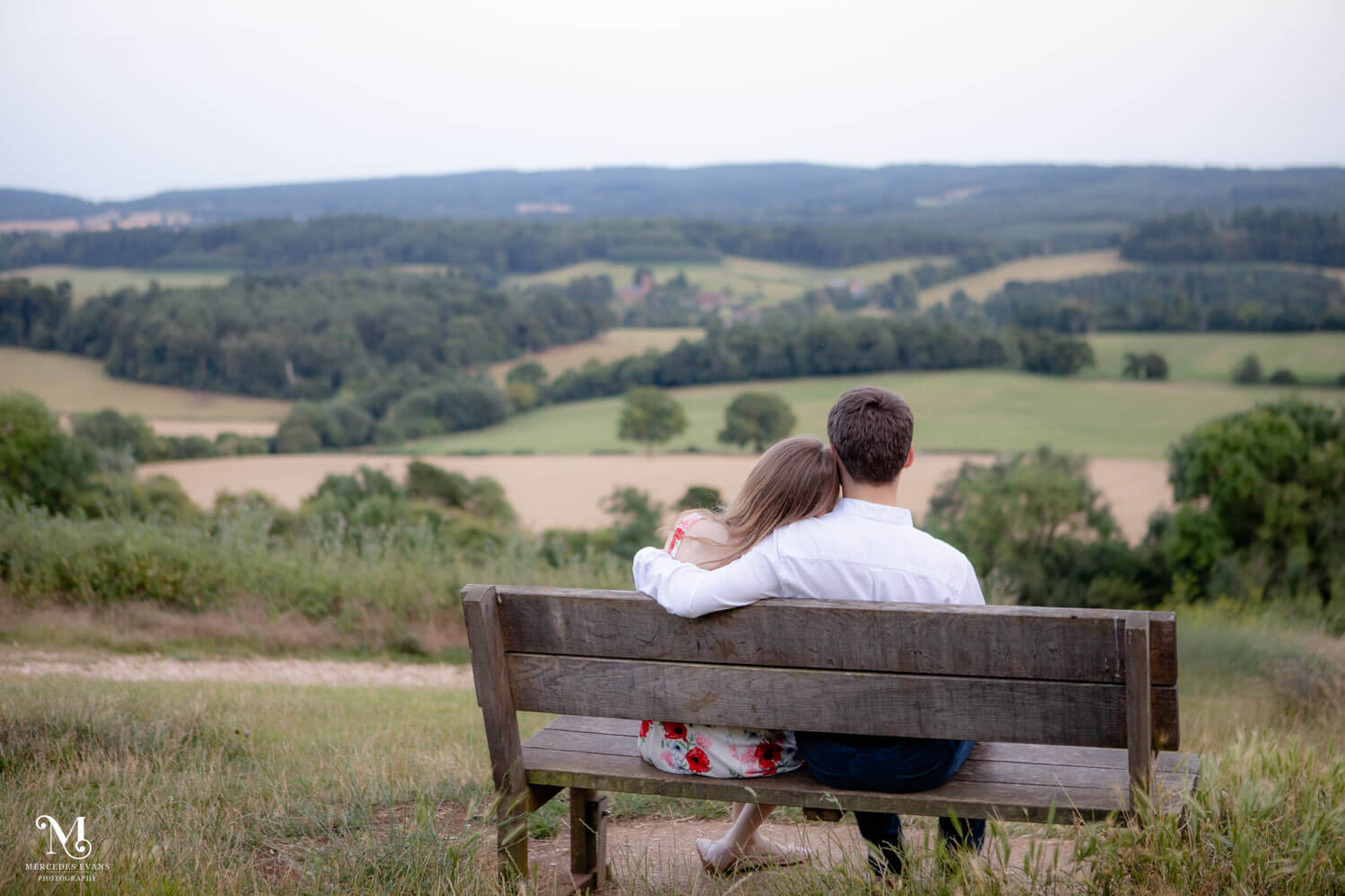 young woman rests her head on young man's shoulder. as they sit on a bench overlooking the Surrey Hills