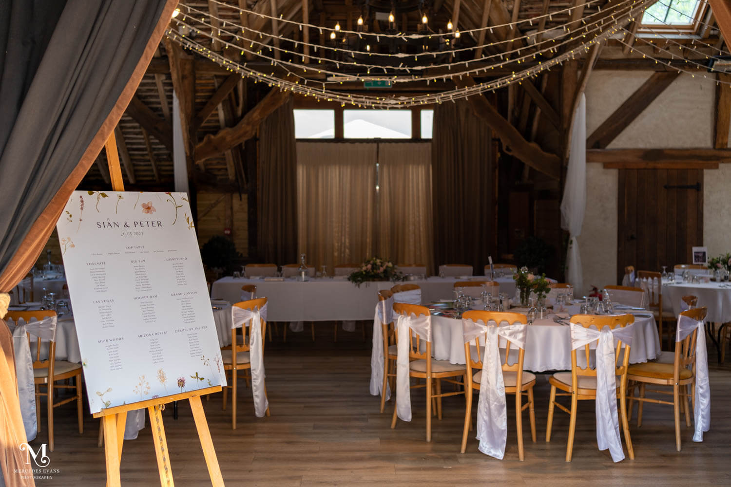 table seating plan stands by the barn doorway, with the tables set for the wedding breakfast behind