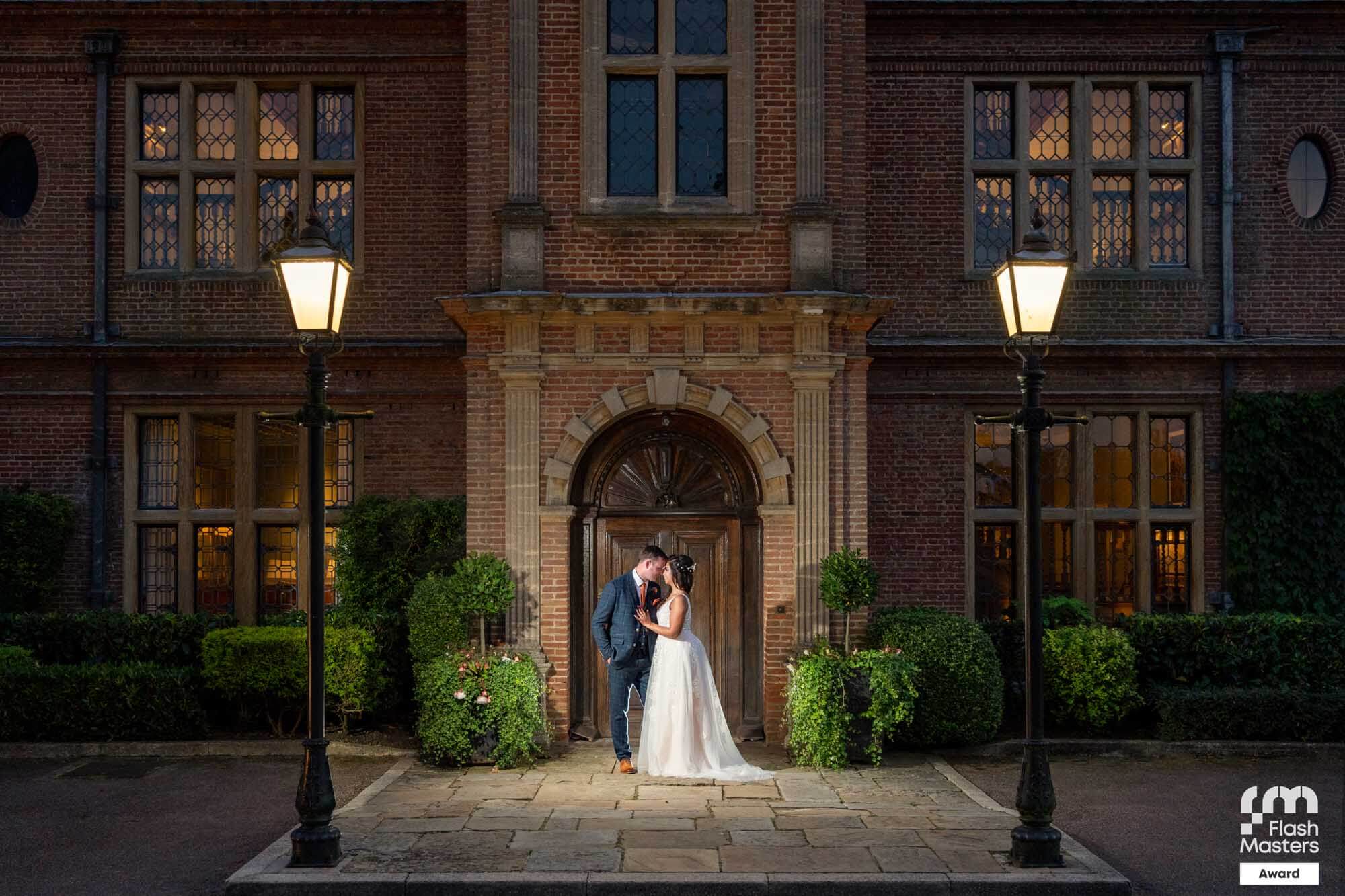 bride and groom stand close together in front of the wooden door of a mansion house in the evening, there are lit lamp posts either side of them