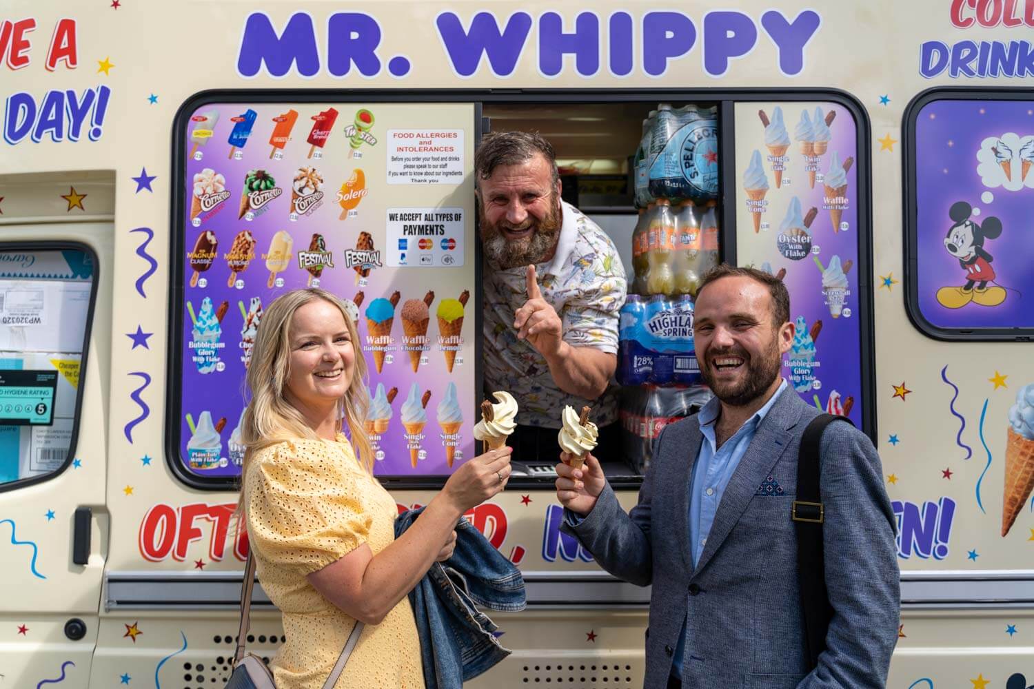 man and woman laugh as they hold their ice creams in front of the mr whippy van, the smiling ice cream seller holds up his index finger