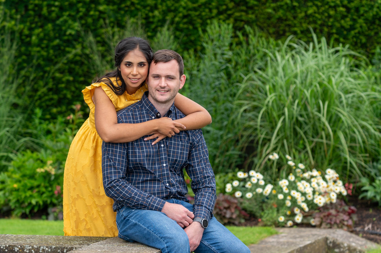 woman in yellow dress hugs her fiancé as he sits on a stone wall in a beautiful garden