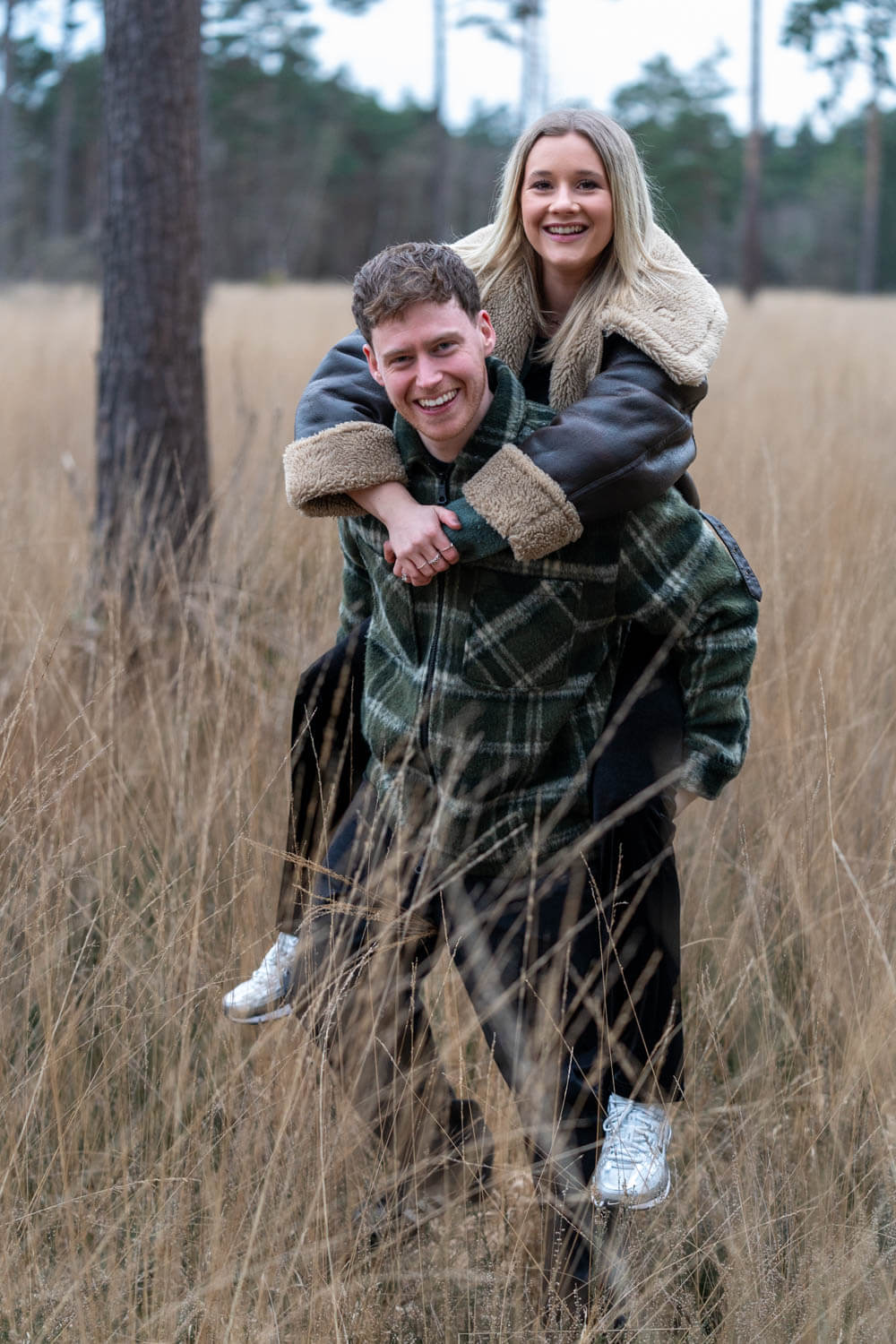 young man gives his fiancee a piggy back in the long grass, both of them are smiling