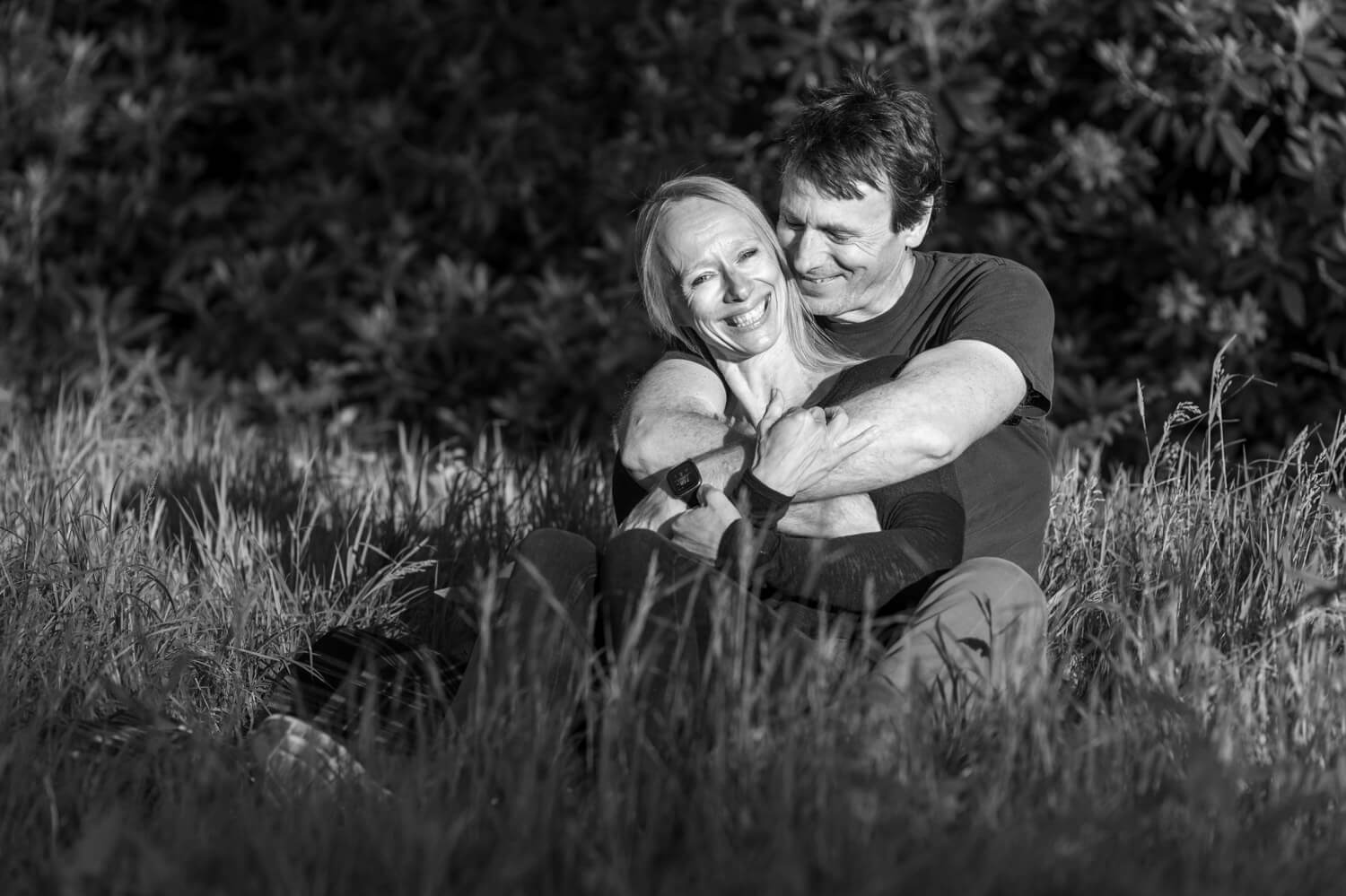 black and white of man hugging a smiling woman as they sit in long grass