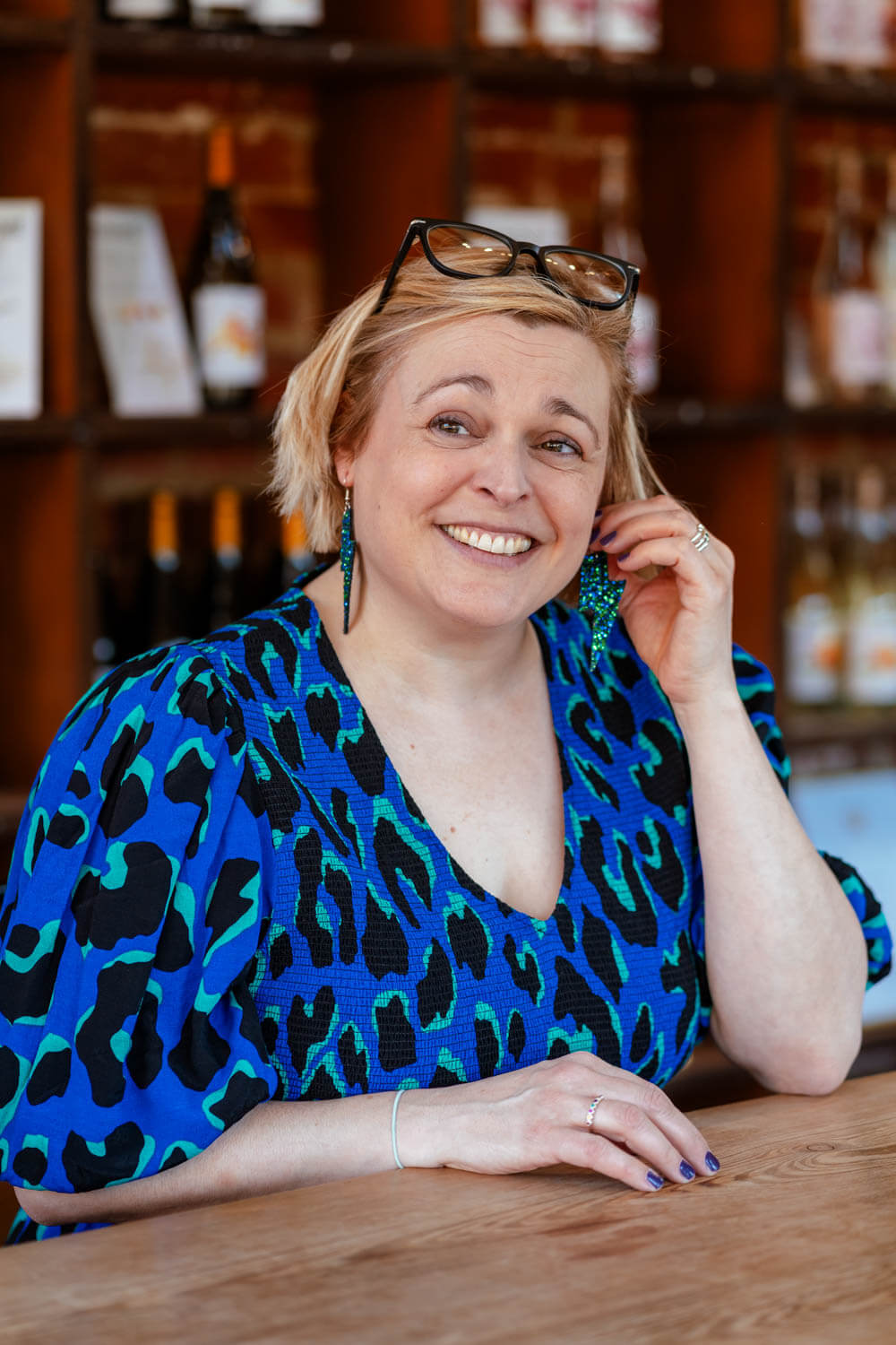 smiling woman in a bright blue tiger print dress plays with her earring, her glasses are perched on top of her short blonde bob