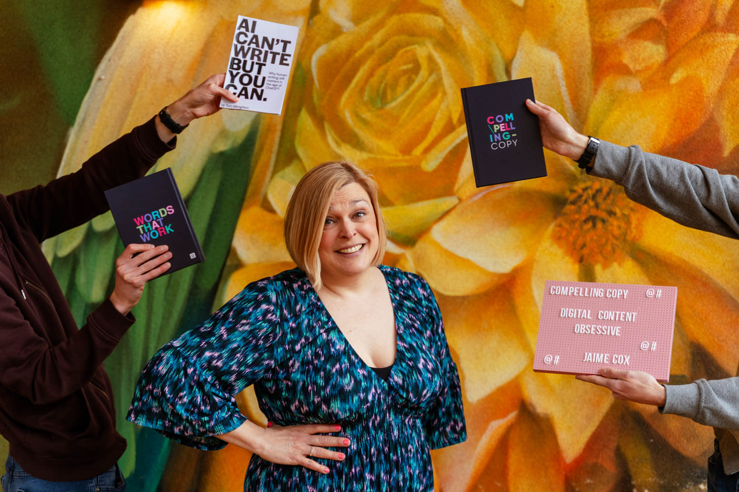 jaime cox female copy writer stands in front of a mural of yellow flowers, hands on either side of her hold up books and a sign
