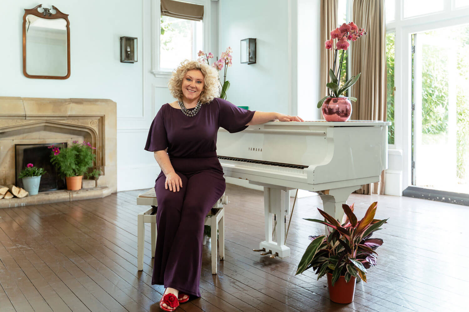female pianist wearing a dark plum jumpsuit sits on the piano stool next to the white grand piano, there are pink orchids on the top of the piano
