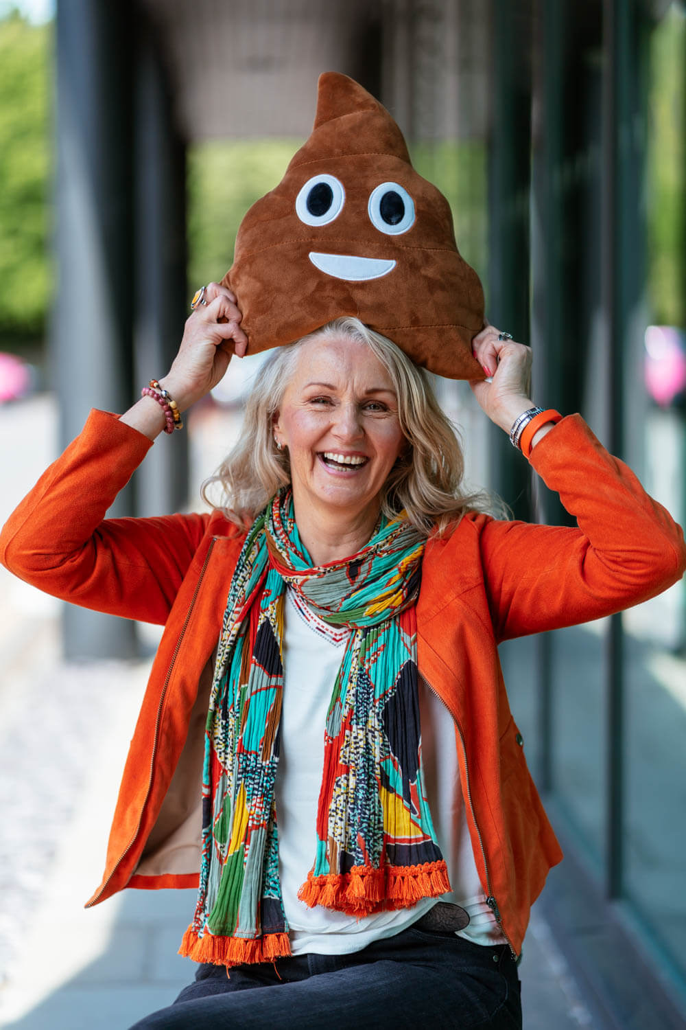 laughing female in a bright orange jacket and multi coloured scarf balances a toy poo cushion on her head