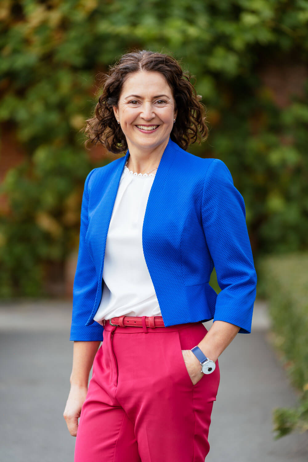 smiling lady wearing white top, electric blue jacket and pink trousers stands in front of a leafy background