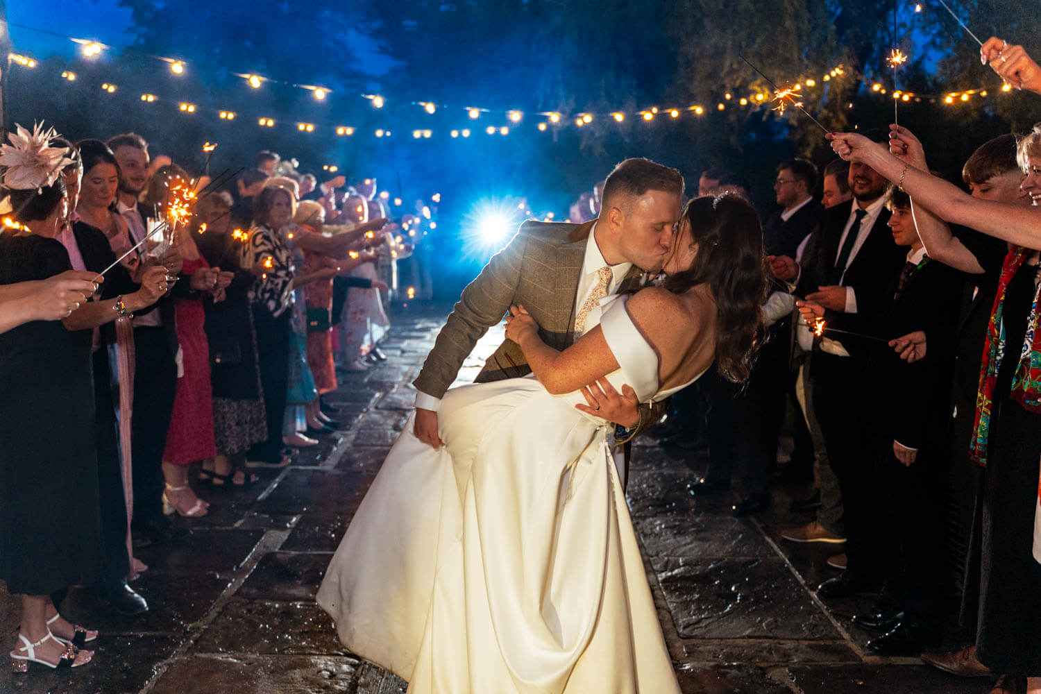 the bride and groom do a dip and kiss as their wedding guests stand watching and waving sparklers on the patio at cain manor