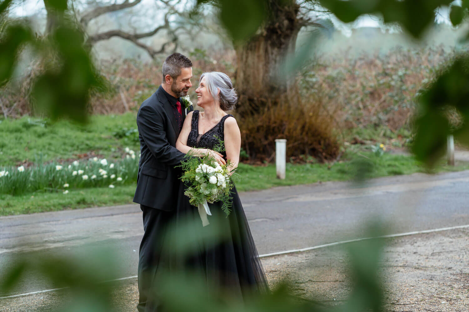 the bride and groom laugh as they stand close together, there is green foliage in the foreground and daffodils behind the couple