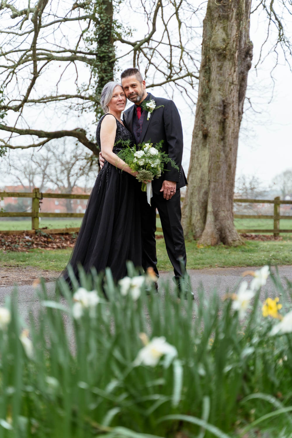 bride in a black wedding dress snuggles up close with her new husband, daffodils are in the foreground