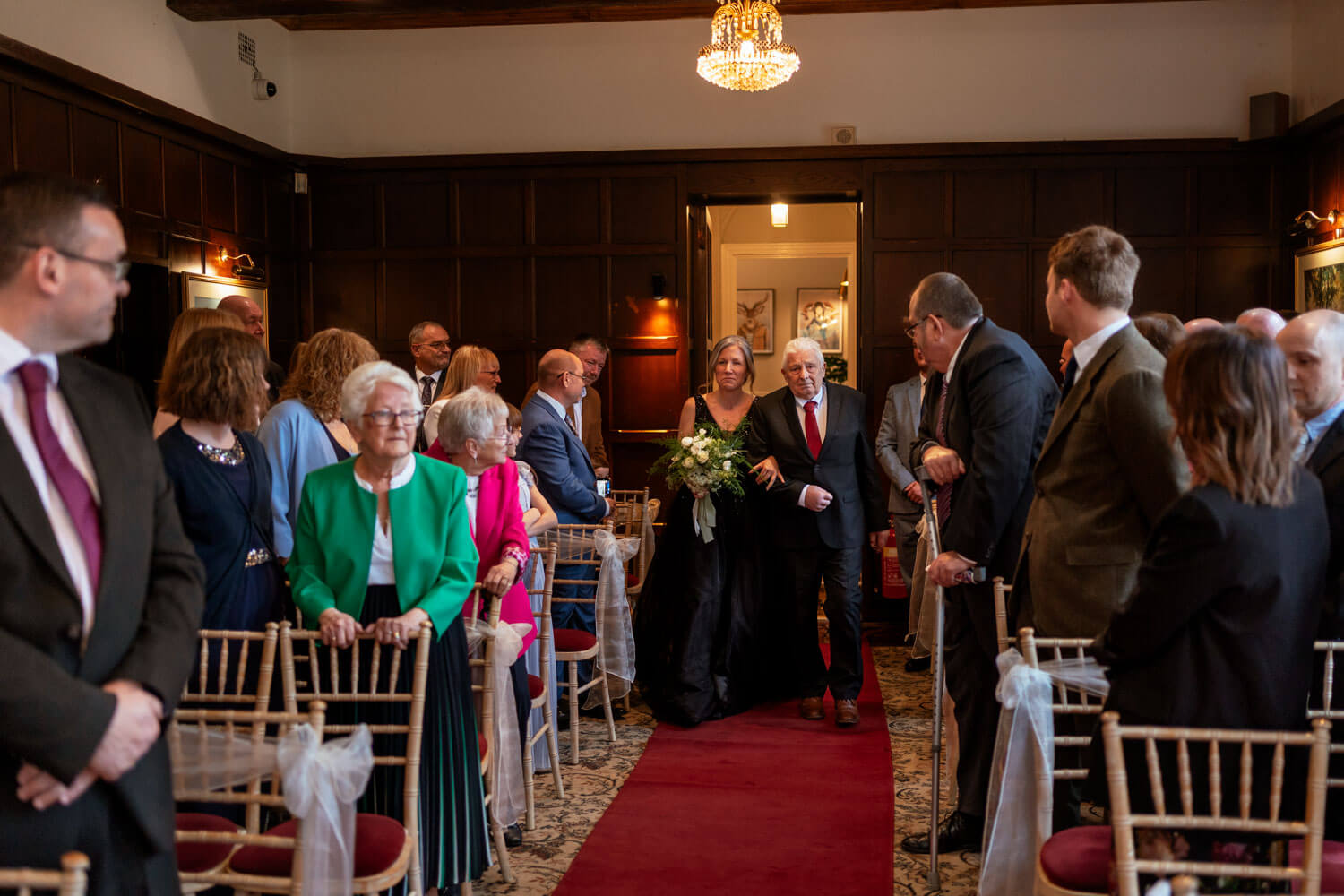 the bride and her father walk down the aisle of the Oak room at Cantley House, the bride wears a black dress and carries a bouquet of white and green florals
