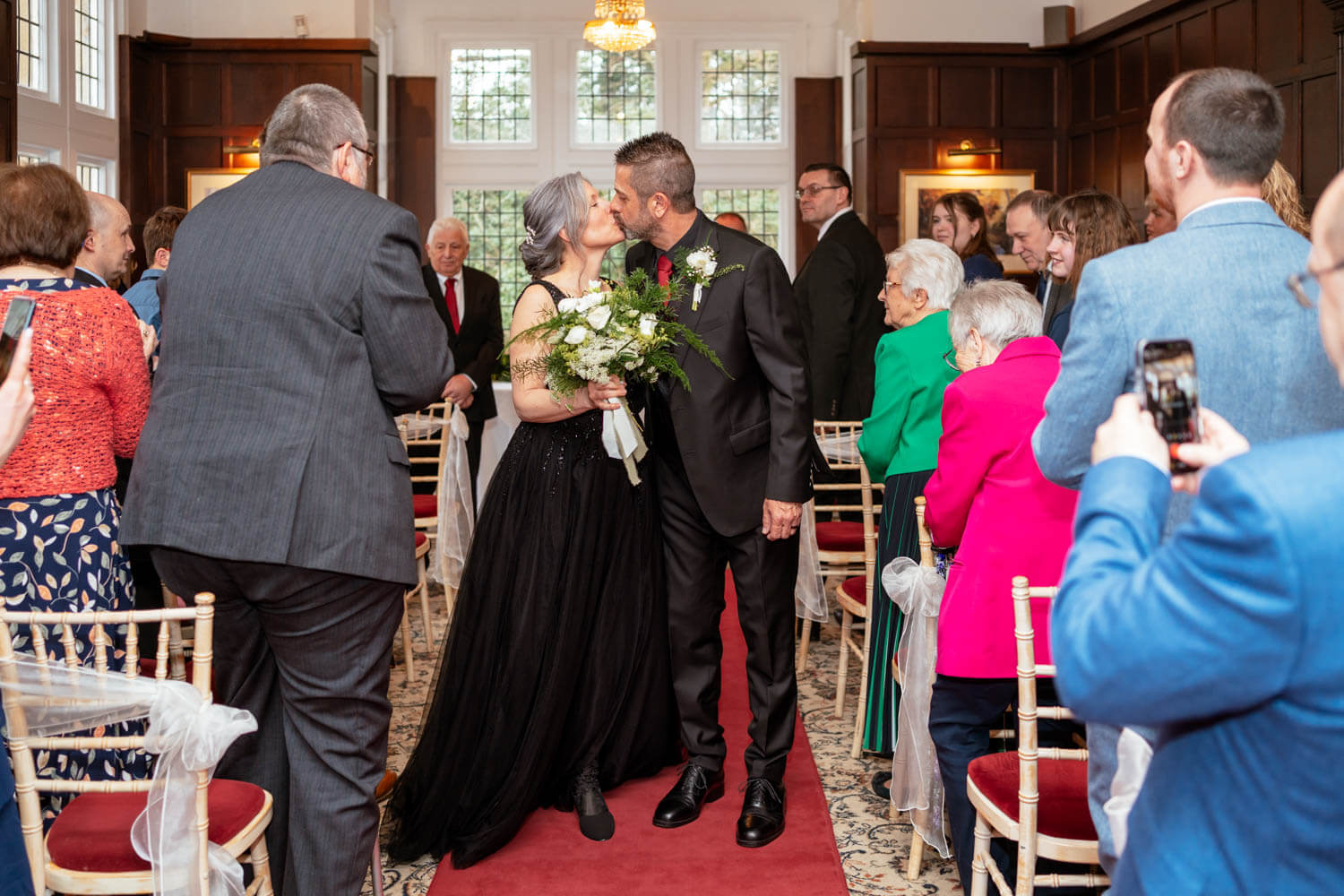 the bride and groom kiss half way down the aisle, as guests watch and snap photos on their phones