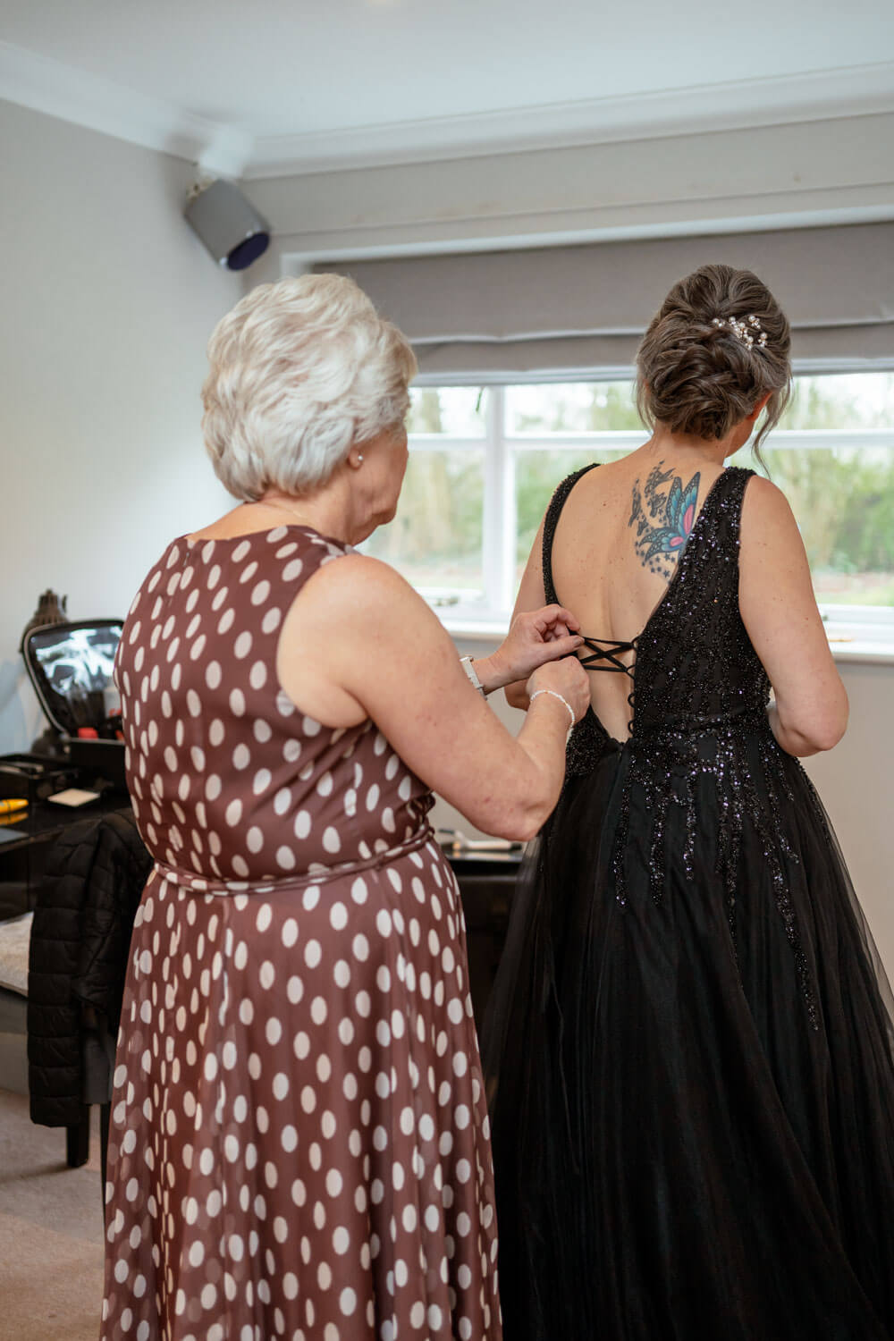 mother of the bride in a brown and white spotted dress ties the ribbons on the back of the bride’s black sparkly wedding dress