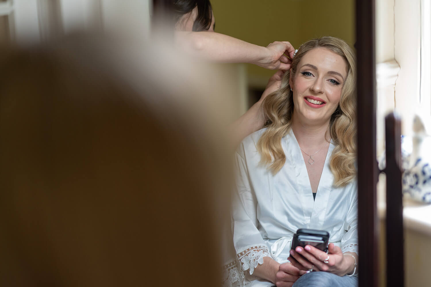 the smiling bride looks in the mirror as the hairdresser fixes her hair