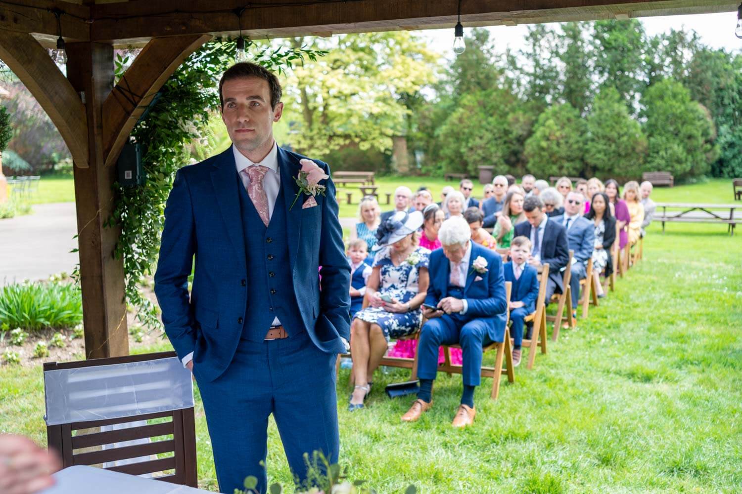 the groom stands at the top of the aisle under the pavilion, waiting for the entrance of the bridal party
