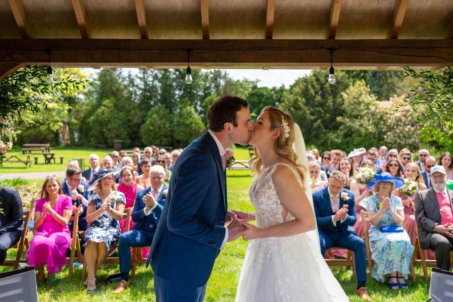 the bride and groom enjoy a first kiss during their wedding ceremony under the Pavilion at Herons Farm, in the background wedding guests applaud