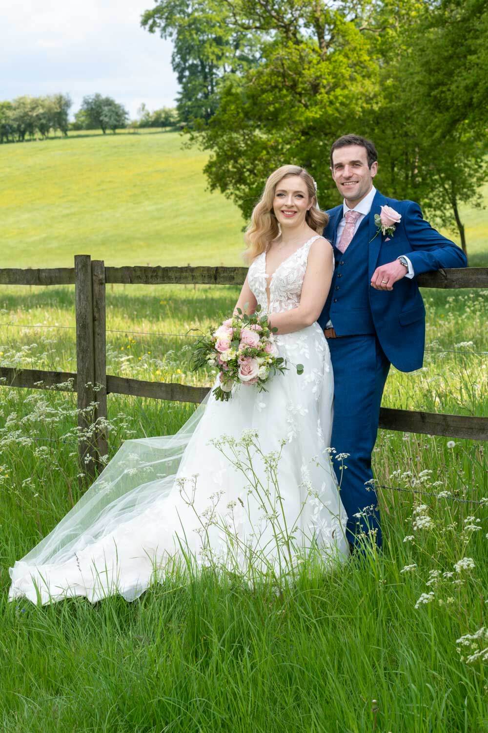 the groom leans against the wooden fence with his bride in front of him during their couple photos at Herons Farm