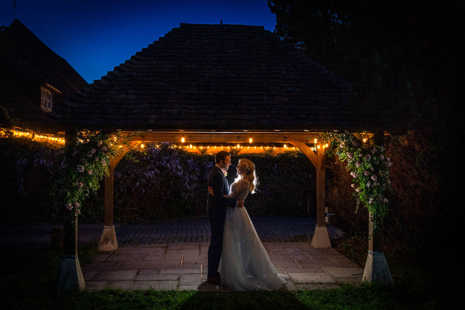 backlit bride and groom stand under a wooden pavilion decorated with fairy lights and flowers at Herons Farm Berkshire