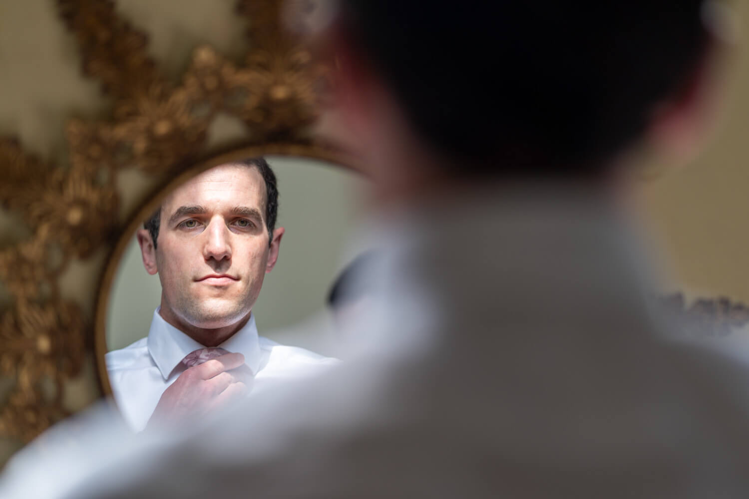 the groom looks in the mirror as he ties his tie